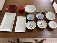 Photo showing the full set of bowls and plates arranged on a wooden table including rectangular sushi plates, lacquered bowls, white and blue patterned bowls, and small cups.