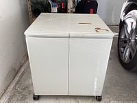 Front view of the white metal storage cabinet with doors and castors, showing closed doors and scuffed top surface.