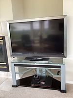 Sony Bravia 46-inch flat screen TV sitting on a metal and glass TV stand in a living room setting. Black screen with silver bezel frame visible, TV stand has clear glass top and bottom shelves with metallic silver legs. Remote control and cables on bottom shelf.