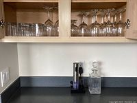 Wide photo showing the Secura electric wine opener in charging base and square glass decanter on counter beneath mounted cupboard.