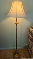 Full view of the brass tone metal floor lamp showing the white fabric lampshade and base with claw feet against a light blue wall.