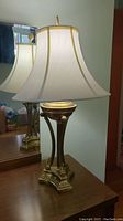 Side view of brass tone table lamp with white shade on wooden surface next to mirror.