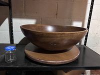 Large dark brown wooden salad bowl on wooden turntable base with small jar next to it for scale. Wall background.