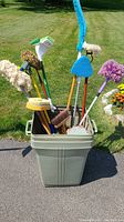 Photo showing large green garbage can filled with assorted long handle cleaning tools, including mops, brushes, scrubbers, and brooms.