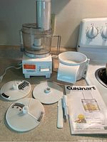Full view of Cuisinart Handy-Prep 3 Cup Food Processor with clear bowl, lid, three different blades/discs, scraper tool, and instruction booklet on kitchen counter.