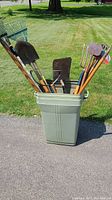 Full view of the green plastic garbage can filled with assorted long handle garden tools on paved surface with grassy lawn background.