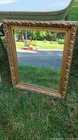 Front view of the mirror showing gold ornate frame and clear reflective glass, standing on grass outdoors.