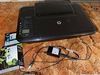 Closed view of HP DeskJet 3050 printer showing control panel, power button, and HP logo. Two unopened HP 61 ink cartridges and a power adapter with cord placed next to the printer on carpeted floor.