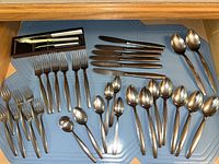 Shows the full assortment of flatware including forks, knives, various spoons and a boxed set of cutting utensils on a blue textured surface.