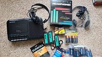 Sony TCM-818 Cassette Corder with wired headphones, battery holders, multiple packs of Duracell and Energizer batteries, and one boxed set of headphones on carpet.