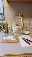 Overview of miscellaneous kitchen items on countertop including mugs, salt and pepper shakers, cutting utensils, paper towel holder, ridged white plate, and Pampered Chef butternut bar board in package.