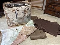 The photo shows a packaged neutral floral comforter set with embroidered flower details and brown and cream colors. The set includes pillow shams, pillow covers, and a brown bed skirt. Next to the package are various curtains and a dragonfly themed shower curtain all in neutral brown tones.
