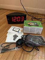 Photo of Garmin Nuvi 56 LM GPS box, mount, USB cable, manuals on floor next to a large red LED alarm clock radio showing time 12:03.