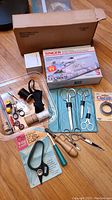 Full lot overview showing sewing machine box, scissor set, thread spools, cutting edge scissors packaging, wooden handled seam ripper, and other small sewing accessories.