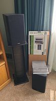 Two Paradigm OM-100 speakers, one on an assembled Ossullivan CD Tower stand, the other speaker on floor next to a boxed Ossullivan CD Tower stand and Paradigm manuals.