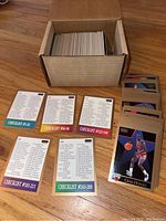 Photo showing a box containing stacks of basketball cards with six checklist cards laid out in front numbered in various ranges corresponding to the card numbers included in the set.