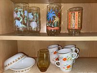Full set of vintage Corelle or Corning glassware and dinnerware items displayed in wooden cabinet shelf.