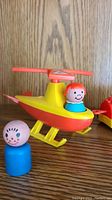 Close-up photo of yellow and red vintage Fisher Price helicopter with orange rotor blade and a pegperson figure with red hair in cockpit with a blue-bodied pegperson figure nearby.
