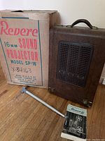 Vintage Revere 16mm Sound Projector box, carrying case with built-in speaker, extension tube, and original manual on wood floor.