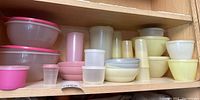 Wide view of kitchen cabinet shelf holding varied vintage Tupperware storage bowls and containers in pink, cream, white, and yellow tones.