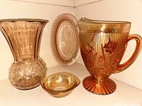 Four vintage glassware items displayed together on shelf: pink fluted vase, floral iridescent pitcher, small iridescent bowl, oval relish/celery dish