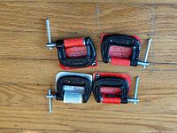 Four 1-inch C clamps laid on wooden surface showing size and condition, including visible red protective grips and metal screws.
