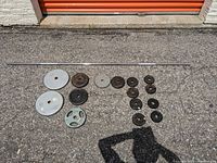 Full view of all weight plates and the barbell laid out on pavement outside near orange garage door. Barbell is a long steel bar, plates arrayed by size and weight.