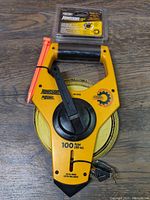 Yellow Johnson open reel measuring tape with black handle and orange latch on wood floor, measuring tape wound on reel visible