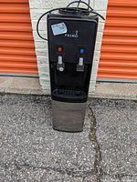 Full front view of the water dispenser showing power cord, body, and base on pavement outside storage lockers.