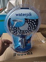 Front view of boxed Waterpik Rainfall Showerhead in clear plastic packaging showing product and branding.