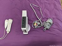 Top-down view showing two white Wii Remote controllers, the vertically positioned white Wii console on its stand, and bundled AV and power cables on a purple background.