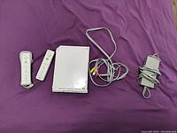 View of Nintendo Wii console with two controllers, AV cable, and power adapter on purple background.