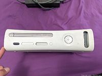 Front view of white Xbox 360 console showing disc tray and power button held against purple background.
