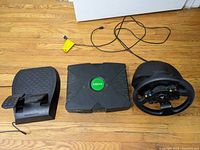 Top view of the Xbox console, steering wheel with buttons, and pedal set on a wooden floor.