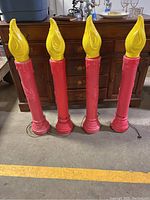 Four red and yellow blow mold electric lawn candles with wired cords on the ground in front of wooden dresser.