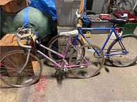 Both bikes positioned side-by-side in a garage against a wooden block and covered storage, showing pink Murray and blue General bikes.