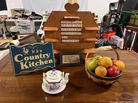 Full view of the lot showing wooden perpetual calendar, Country Kitchen sign, fruit basket, and metal teapot on wooden surface with workshop background.