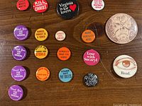 Multiple novelty and business related buttons including humorous and political quotes in various colors and sizes photographed on wooden surface.