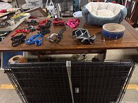 Dog accessories including small dog bed, large ceramic bowl, multiple dog leashes and harnesses on a wood table, with large black wire dog crate folded and leaning against table