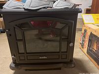 Front view of Duraflame electric stove heater showing black textured finish, glass door with decorative framing, and handle on right side. Cardboard box with product info visible nearby.