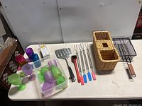 Wide view of all items on a table including grill tools, colored drinking cups, plastic wine glasses in a container, and woven utensil basket.