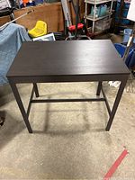 Front diagonal view of brown IKEA bar table showing solid tabletop and four legs connected by support bars, placed in a warehouse environment.