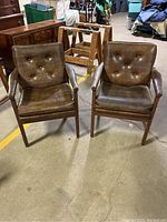 Front view of two Mid-Century Modern style accent chairs with wooden frames and brown leather upholstery sitting on concrete floor among other furniture and items.