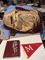 Varsity jacket with inside lining "Milton" embroidery, laid out flat showing maroon and gold colors with striped cuffs; 1977 yearbook and red pennant placed below