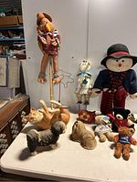 Full view of the lot showing all stuffed animals, ceramic cat figurine, snowman figurine, teddy bears and dolls on table.