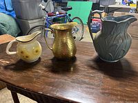 Three vintage decorative pitchers displayed on a wooden table in a storage area with various items in the background