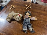 Wooden marionette boy puppet lying flat on a table alongside a plush dog-like doll with yarn hair and red bows.