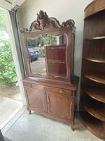 Full front view of the antique wooden cabinet with two sections, top hutch with decorative carved crest and mirrored door, bottom base cabinet with drawer and doors.