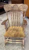 Front view of antique wooden rocking chair showing carved backrest with shell and scroll motifs, spindle back, and worn wooden seat.