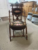 Front view of antique chair showing floral needlepoint upholstery on seat and oval backrest, carved wooden frame with turned legs and ornamental finials.